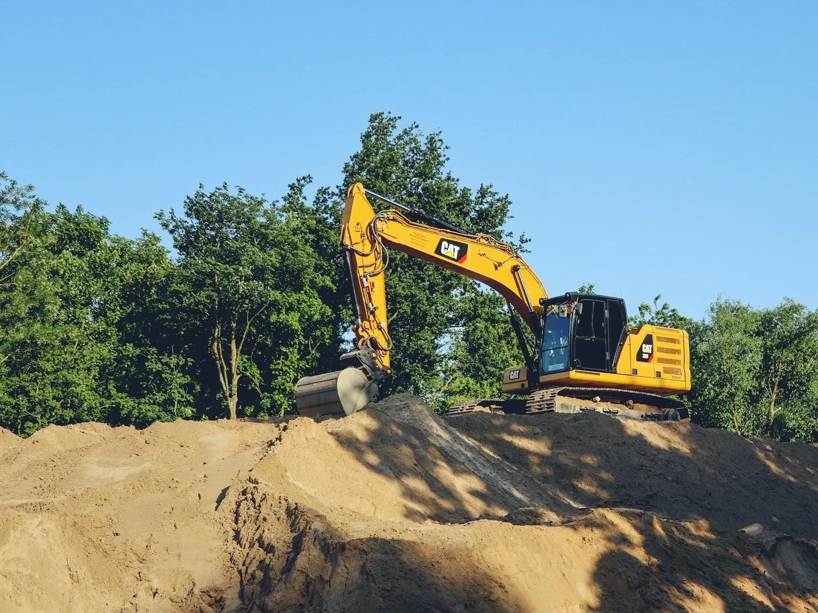 Cat excavator on a mound of dirt with a line of trees behind