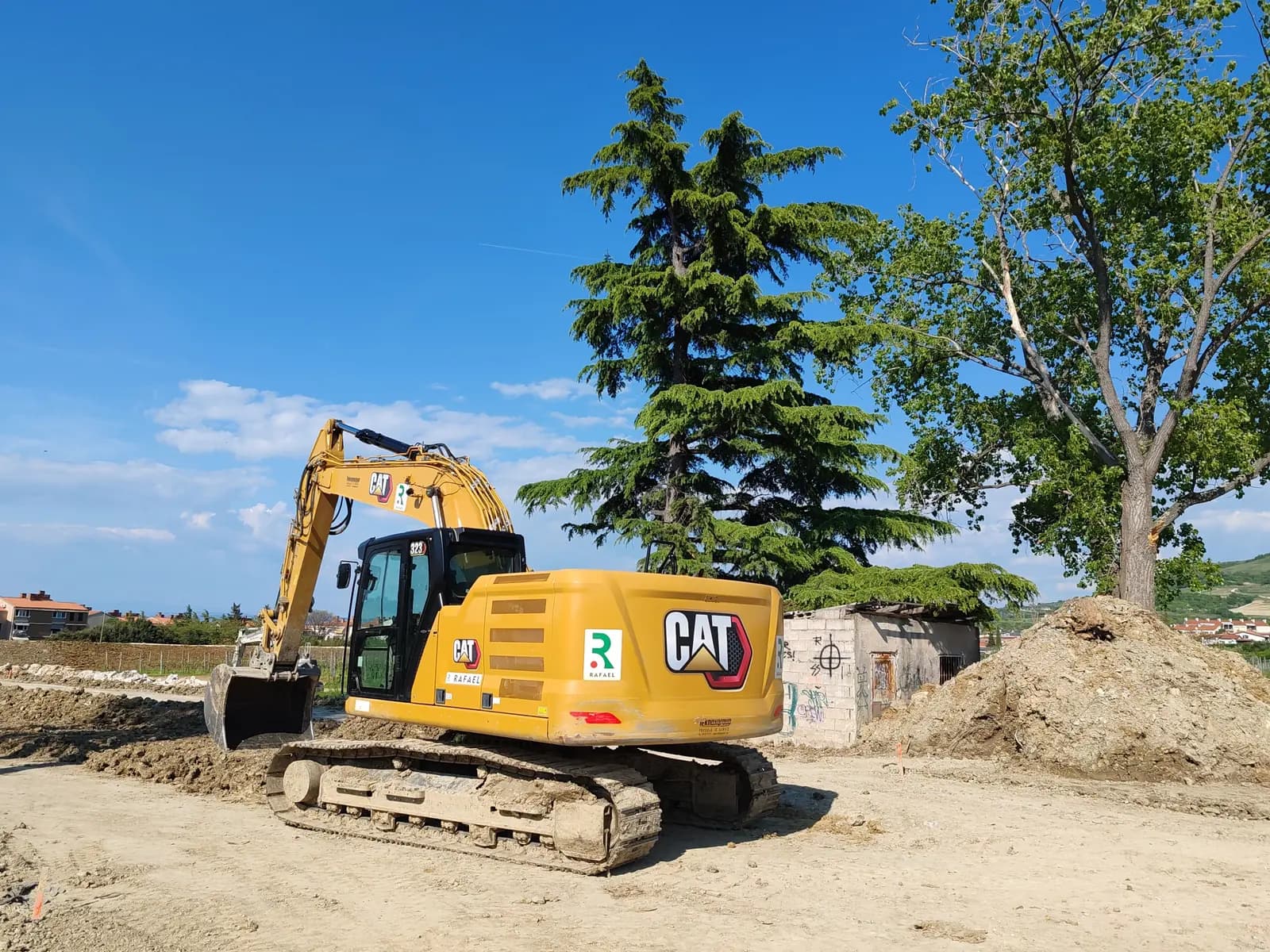 Cat excavator on a dirt work site beside tall evergreen and shade trees