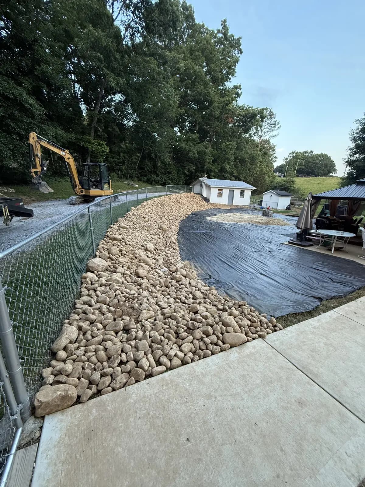 Rock-lined drainage swale over landscape fabric along a fence, with a mini excavator on site, by Superior Land Management