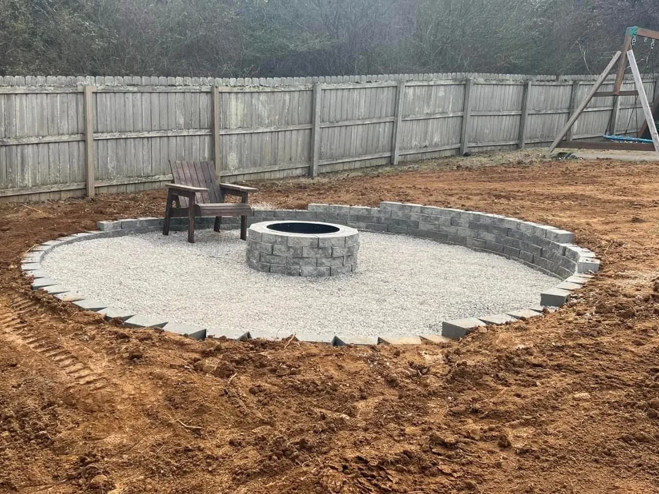 Circular gravel fire pit patio with a stone block border and fire ring on freshly graded red clay